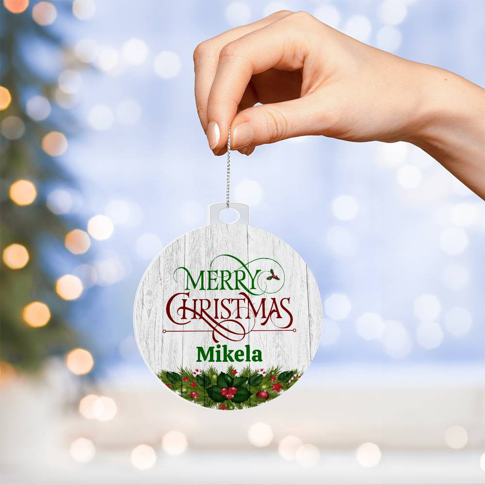 A hand holds the AnywherePOD Personalized Name Christmas Ornament, showcasing "Merry Christmas Mikela" inscribed on it. This custom keepsake is adorned with green and red holly against a snowy backdrop, complementing the blurred holiday lights and tree in the background beautifully.