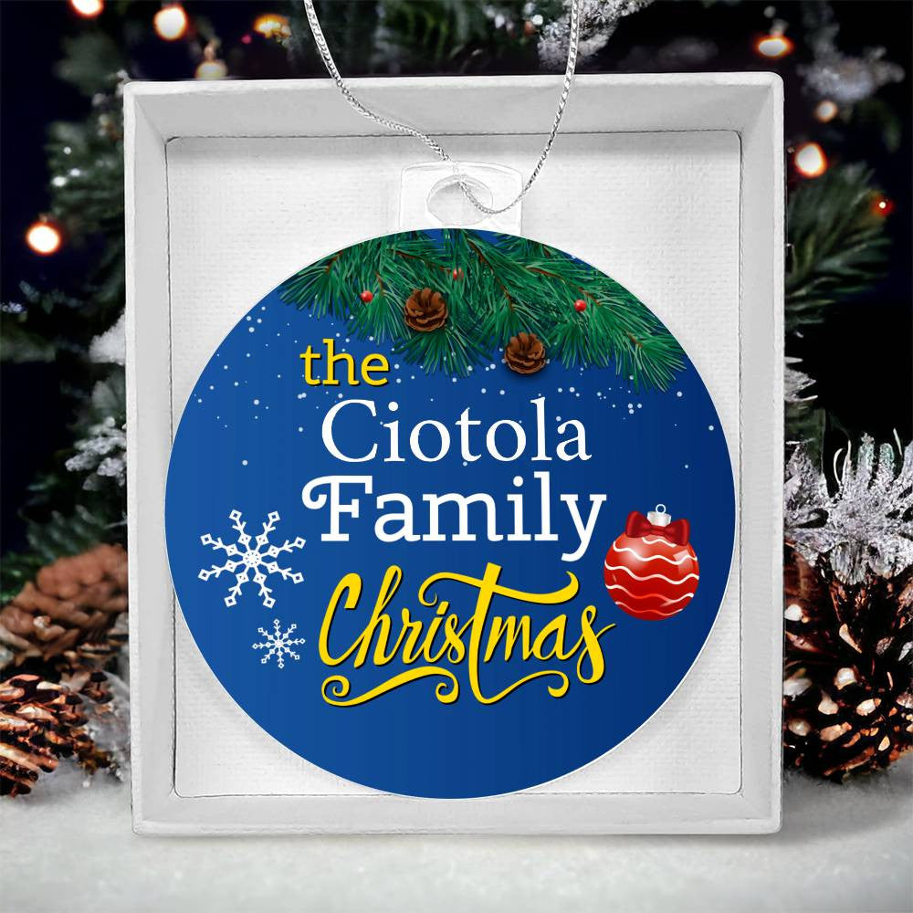 The AnywherePOD "Create Cherished Memories with a Personalized Family Name Christmas Ornament" features the Ciotola Family Christmas message nestled in a gift box. Crafted from high-quality acrylic, it is designed with pine branches, pinecones, snowflakes, and a red bauble. A beautifully decorated Christmas tree and additional pinecones in the background enhance the charm of this unique keepsake.