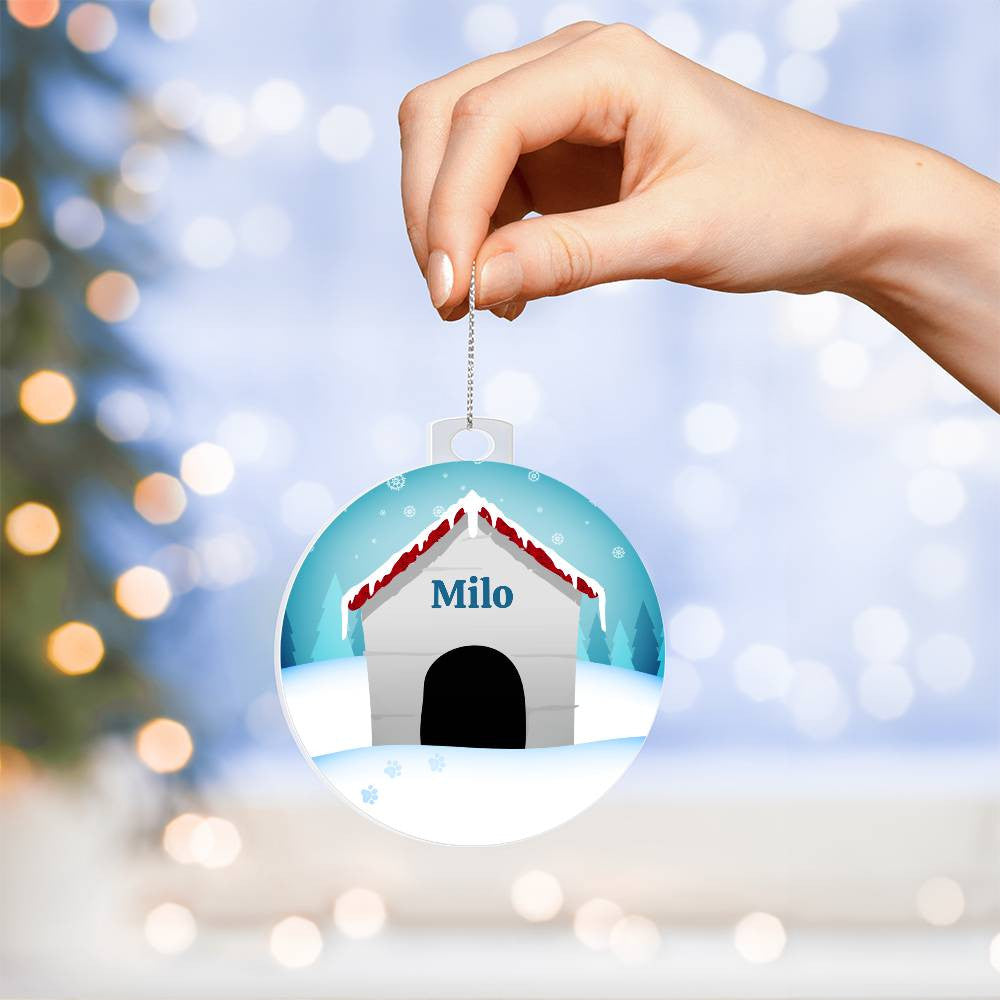 A hand displays AnywherePOD's "Honor Your Pup This Holiday with a Personalized Dog Name Christmas Ornament," featuring an acrylic ornament with a doghouse adorned with a red roof and personalized with the name "Milo." The backdrop includes a winter scene accented by softly blurred lights and a glimpse of a Christmas tree on the left, creating an exceptional keepsake for the holiday season.