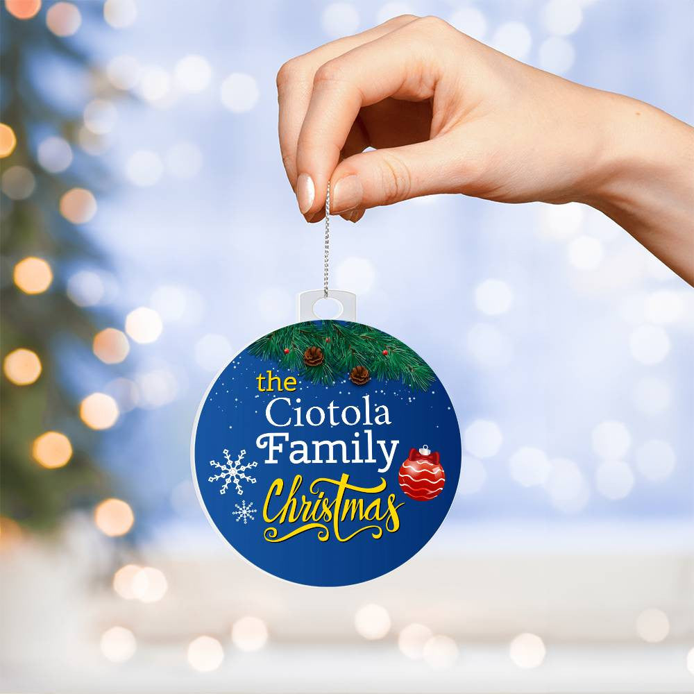 A hand holds the "Create Cherished Memories with a Personalized Family Name Christmas Ornament" by AnywherePOD, crafted from high-quality acrylic and showcasing "The Ciotola Family Christmas" on a blue background adorned with green pine needles and pinecones. Festive designs of a snowflake and red ornament add charm, while blurred lights twinkle softly in the background.