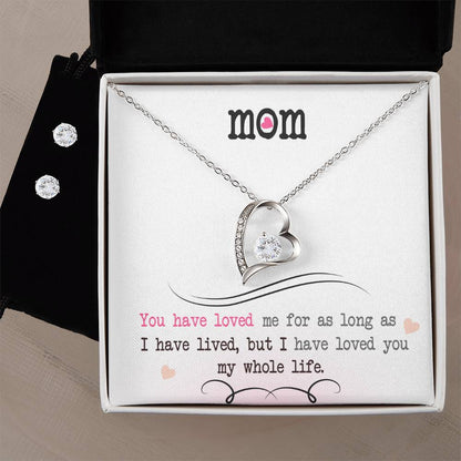 The Forever Love Necklace, branded by ShineOn Fulfillment and featuring a heart-shaped design in 14k White Gold Finish, is elegantly presented in an open jewelry box. The lid displays the word "Mom" adorned with a pink heart motif. Below this, a heartfelt message is printed in black and pink text. Accompanying the necklace inside the box is a pair of Cubic Zirconia Earrings.