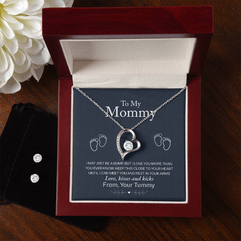 The "To My Mommy, Love From Your Tummy - Forever Love Necklace" by ShineOn Fulfillment, a silver heart-shaped piece with a small diamond and 14k white gold finish, is elegantly presented in an open red velvet jewelry box. The box features a heartfelt message for "Mommy," adorned with tiny footprints. Nearby, you'll find a white flower and a black jewelry pouch.<|vq_7599|>