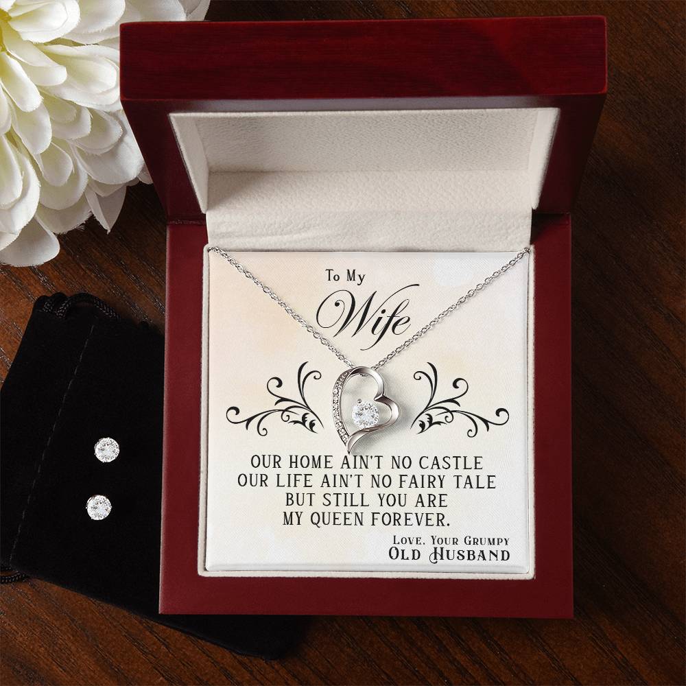 The "To My Wife, You Are My Queen Forever - Forever Love Necklace" by ShineOn Fulfillment, featuring a heart-shaped design with a gemstone and a 14k white gold finish, rests inside a red velvet box accompanied by a heartfelt message to your wife. Meanwhile, a black pouch contains the Cubic Zirconia Earring Set, against the backdrop of white flowers decorating the wooden surface corner.