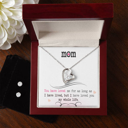 The Forever Love Necklace, crafted by ShineOn Fulfillment in a heart shape with a small stone and a 14k White Gold Finish, is nestled inside a red jewelry box. The box features a heartfelt message dedicated to "mom" about love, decorated with a flower in the corner and accompanied by a black pouch.