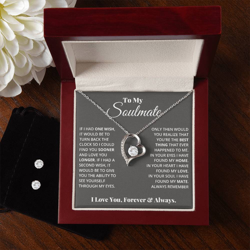 A heart-shaped Forever Love Necklace, with a crystal and 14k white gold finish from ShineOn Fulfillment's "To My Soulmate, In Your Heart I Found My Love" collection, rests in an open red velvet box. The lid of the box is adorned with a sentimental message titled "To My Soulmate." Nearby are white flowers and a Cubic Zirconia Earring Set in a black pouch.