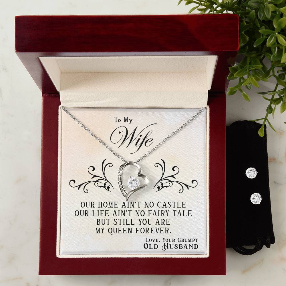 A Forever Love Necklace called "To My Wife, You Are My Queen Forever" from ShineOn Fulfillment, featuring a heart-shaped pendant with a 14k white gold finish, is elegantly displayed in an open wooden jewelry box. The lid of the box displays a heartfelt message to a wife. Accompanying the necklace, a Cubic Zirconia Earring Set rests on a black pouch nearby, with a small plant beautifully positioned in the background.