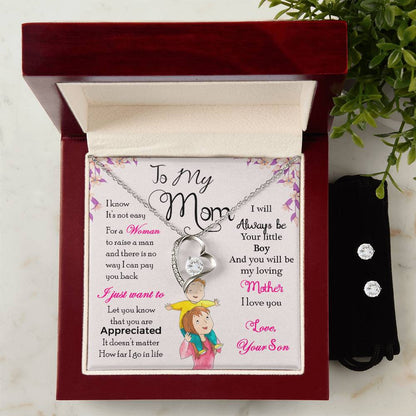 The "To My Mom, I Will Always Be Your Little Boy - Forever Love Necklace" by ShineOn Fulfillment features a silver heart and a sparkling gem, beautifully displayed in an open velvet box with a 14k white gold finish. The box contains a heartfelt message from her son, conveying love and gratitude to "Mom." In the background is a vibrant illustration of a woman holding her child.