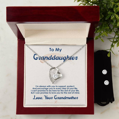 A silver heart-shaped "To My Granddaughter, I Love You For The Rest Of My Life - Forever Love Necklace" from ShineOn Fulfillment, adorned with a small gemstone, beautifully displays its 14k white gold finish in an open box, featuring a heartfelt message from her grandmother. Beside it, a Cubic Zirconia Earring Set sits on a black pouch over the marble surface with greenery.