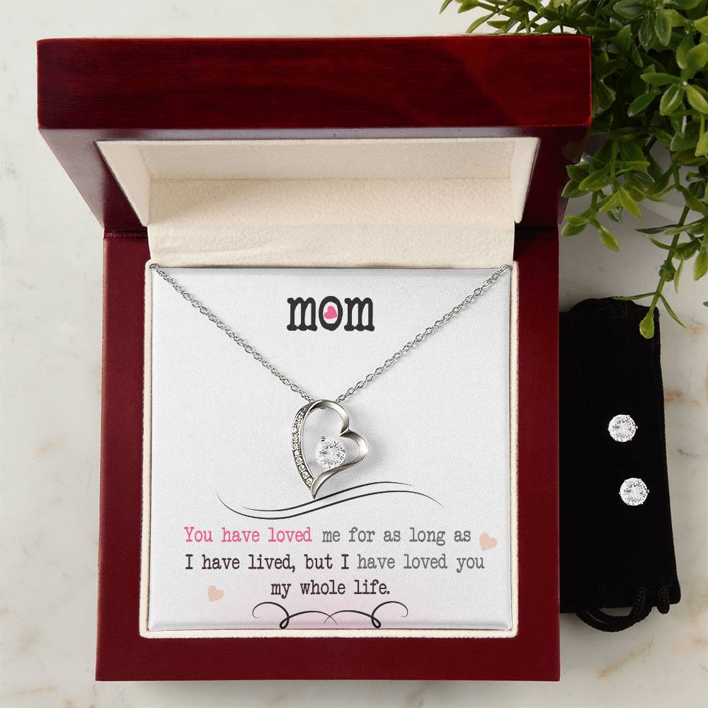 The "To My Mom, I Loved You My Whole Life - Forever Love Necklace" by ShineOn Fulfillment features a heart-shaped pendant with a crystal and is finished in 14k White Gold. It is presented in an open red and white box with the message, "MOM: You have loved me for as long as I have lived, but I have loved you my whole life." Accompanying it is a small black pouch containing a Cubic Zirconia Earring Set.