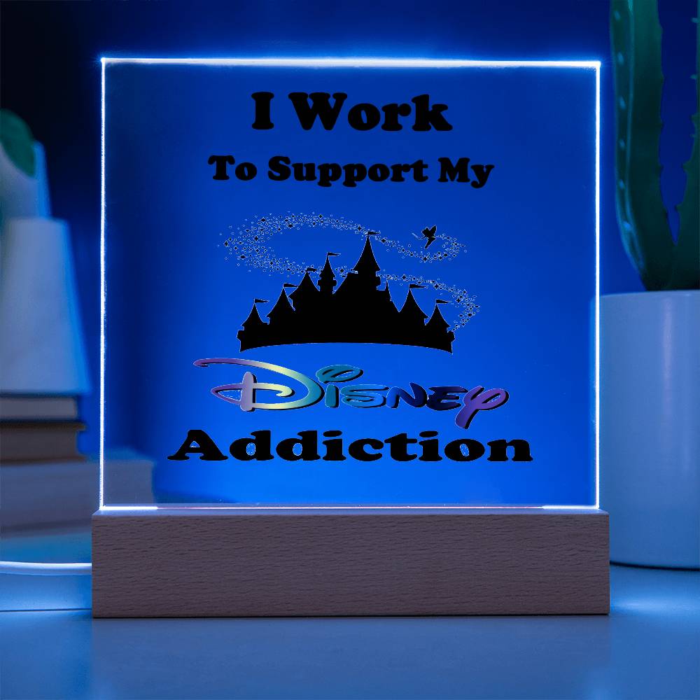 A radiant, enchanting display spells out "I Work To Support My Disney Addiction" in front of a silhouetted castle and sparkles, all set atop a wooden base. Ideal for any Disney enthusiast, the "Disney Obsessed: Where Magic Meets Everyday Life!" acrylic plaque by ShineOn Fulfillment stands against a dark backdrop accompanied by books and a cactus.