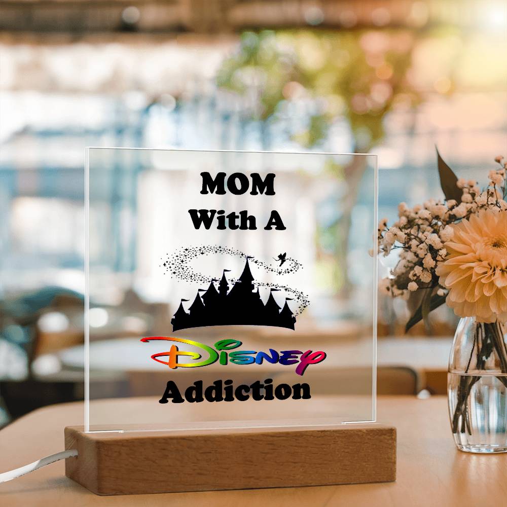 A stylish acrylic plaque from ShineOn Fulfillment, titled "Disney-Obsessed Mom: Where Magic Meets Motherhood!", is set on a wooden base. It features the phrase overlaid with a castle silhouette reminiscent of the Magic Kingdom, complemented by an indoor backdrop showcasing a vase of flowers.