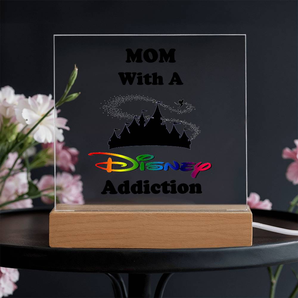 An acrylic plaque titled "Disney-Obsessed Mom: Where Magic Meets Motherhood!" by ShineOn Fulfillment features bold text and a charming castle silhouette. Ideal for any mom who loves Disney, it is beautifully displayed on a wooden base against a softly blurred floral background.