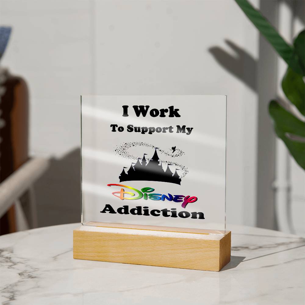 The "Disney Obsessed: Where Magic Meets Everyday Life!" decorative acrylic plaque by ShineOn Fulfillment features the text "I Work to Support My Disney Addiction," complete with a silhouette of the Magic Kingdom castle and a colorful Disney logo. This charming piece, displayed on its wooden stand with a leafy plant in the background, is perfect for any devoted Disney fan.