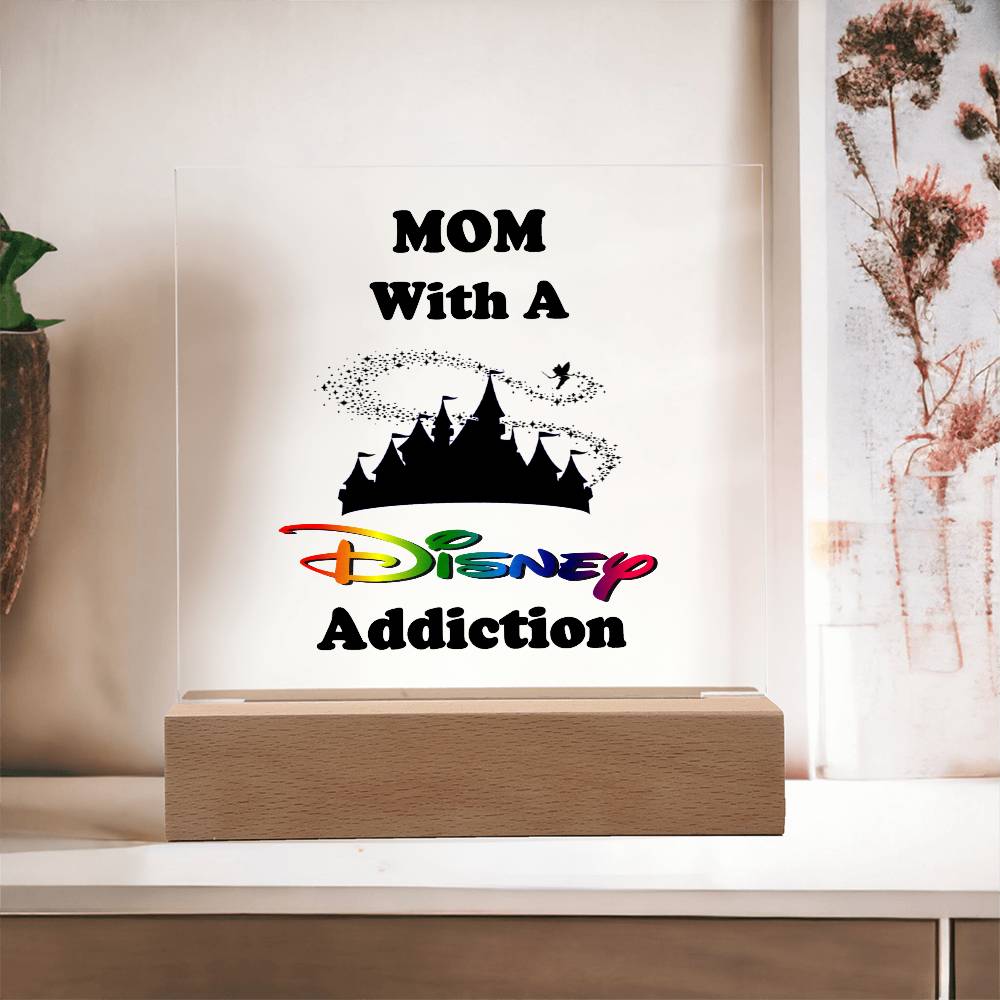 A decorative acrylic plaque on a wooden base from ShineOn Fulfillment displays the phrase "Disney-Obsessed Mom: Where Magic Meets Motherhood!" in colorful letters. The background includes a castle silhouette with a small figure flying above, infusing it with a touch of Mickey magic. The sign is placed on a light-colored surface.