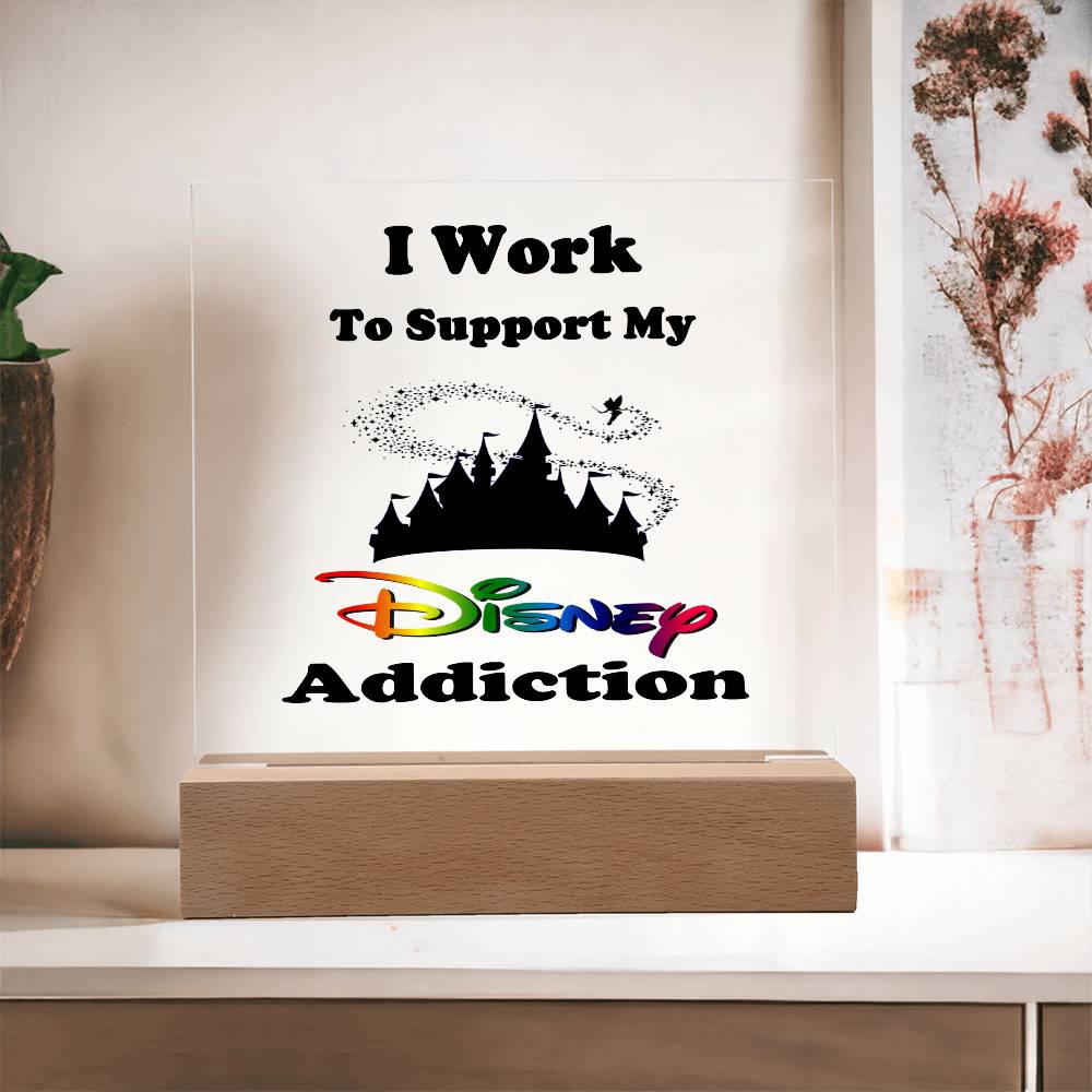 An enchanting plaque from ShineOn Fulfillment, titled "Disney Obsessed: Where Magic Meets Everyday Life!," features the phrase "I Work To Support My Disney Addiction" with "Disney" highlighted in vibrant colors. The plaque is made of acrylic and set on a wooden base, showcasing a silhouette of a castle with Pixie dust from Tinker Bell in the background—ideal for any Disney enthusiast's collection. It stands proudly on a white tabletop against a neutral wall.