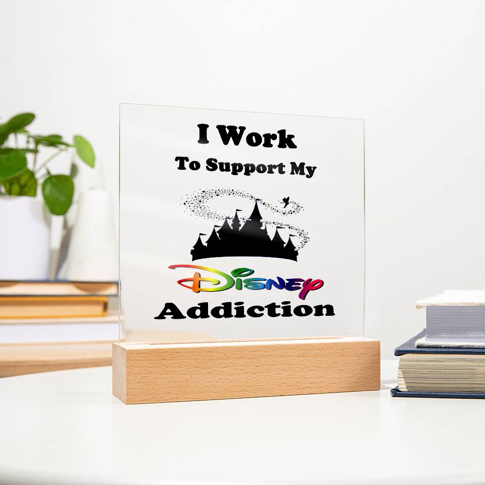 The Disney Obsessed: Where Magic Meets Everyday Life! plaque by ShineOn Fulfillment features vibrant black and colorful lettering declaring "I work to support my Disney addiction," all elegantly displayed on a wooden stand. This enchanting piece graces a white table, nestled beside a stack of books and a potted plant—an ideal addition for any Disney fan's décor.