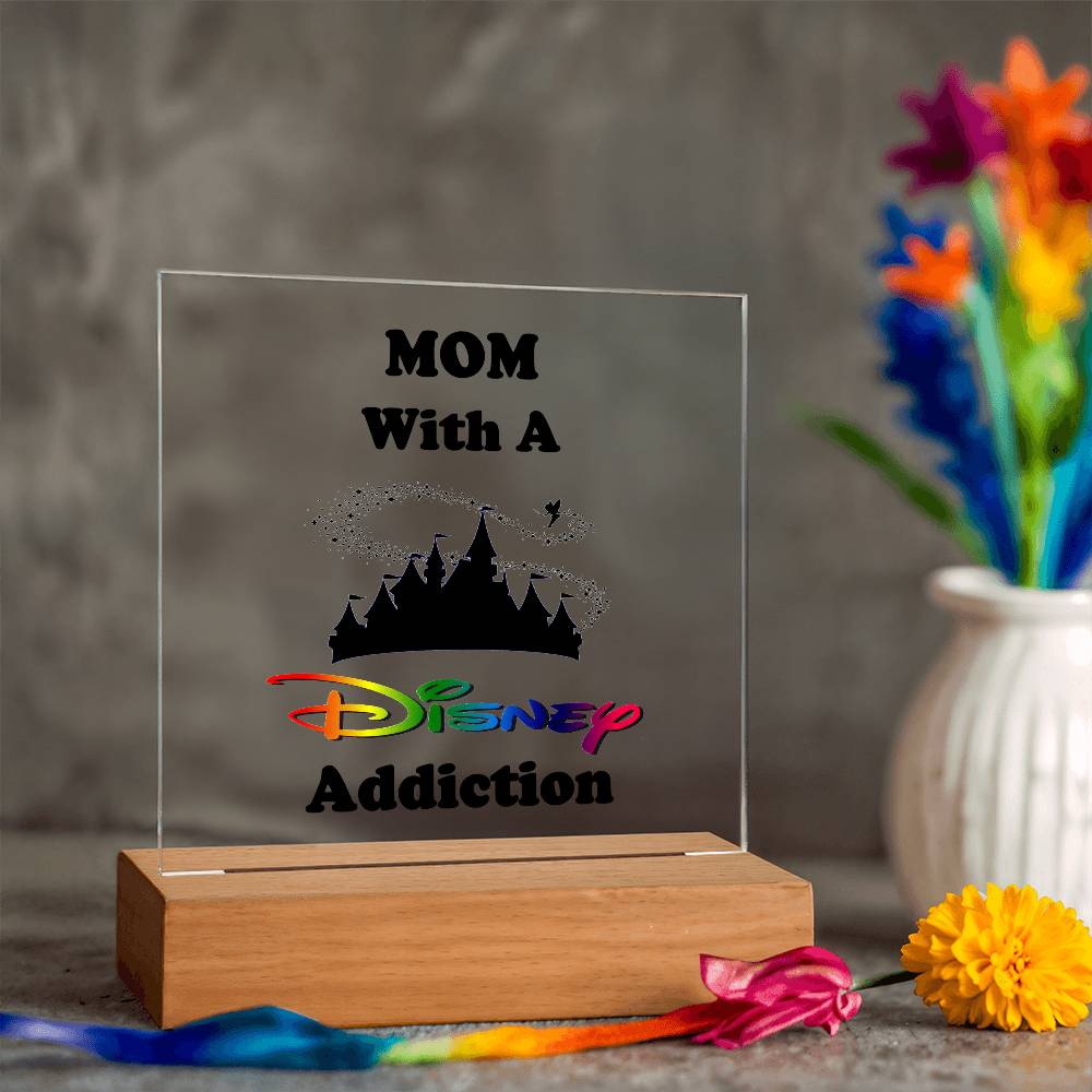 Acrylic plaque on a wooden base reads "Disney-Obsessed Mom: Where Magic Meets Motherhood!" in colorful text that captures the spirit of imagination. In the background, a blurred vase holds vibrant, multicolored flowers. This unique piece is brought to you by ShineOn Fulfillment.