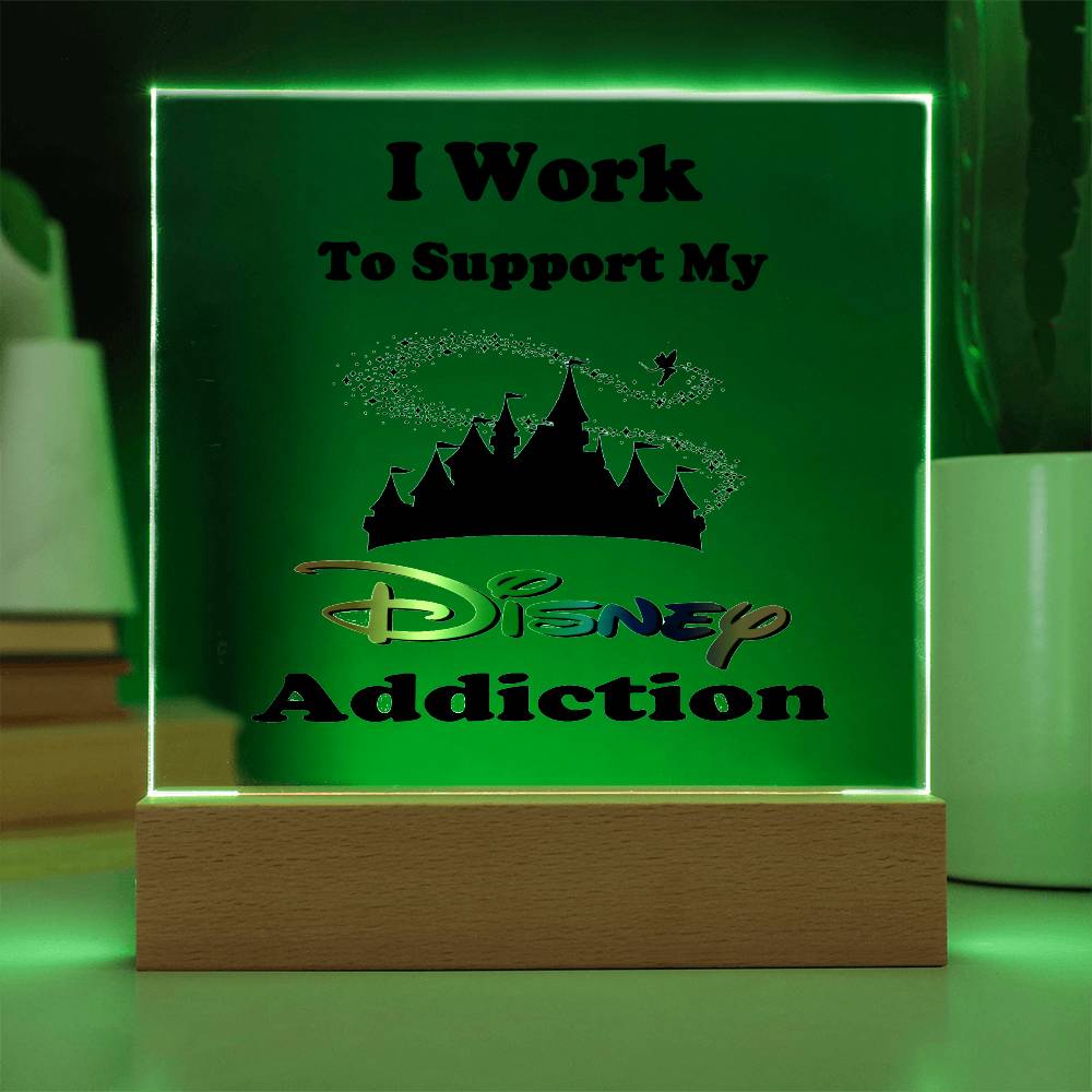 The Disney Obsessed plaque from ShineOn Fulfillment showcases the phrase "I Work To Support My Disney Addiction" alongside a castle silhouette and shimmering accents. This enchanting piece is beautifully illuminated with green light on a wooden base, framed by softly blurred background details.