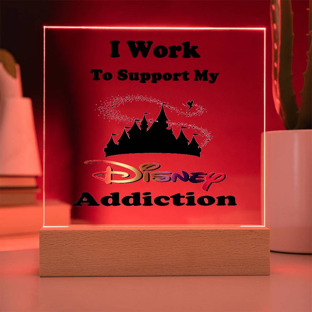 The Disney Obsessed: Where Magic Meets Everyday Life! sign, from ShineOn Fulfillment, features a wooden base and an acrylic silhouette of a castle with the phrase "I Work to Support My Disney Addiction." This enchanting piece stands gracefully against a red backdrop, accentuated by a partially visible plant on the right—ideal for every Disney enthusiast.