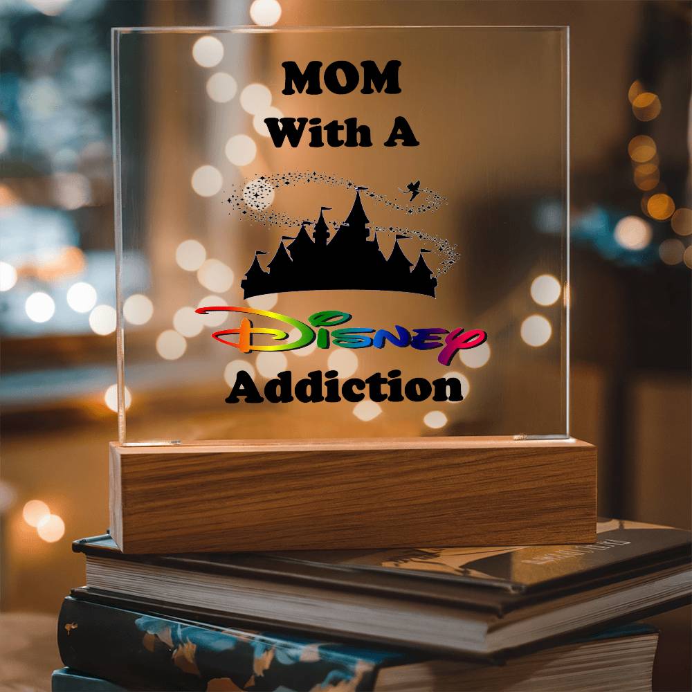 The "Disney-Obsessed Mom: Where Magic Meets Motherhood!" decorative acrylic block by ShineOn Fulfillment features vibrant, iconic letters spelling out "MOM With A Disney Addiction" alongside a castle silhouette. This delightful piece is perfect for any Disney-loving mom and is beautifully displayed on books with twinkling blurred lights in the background.