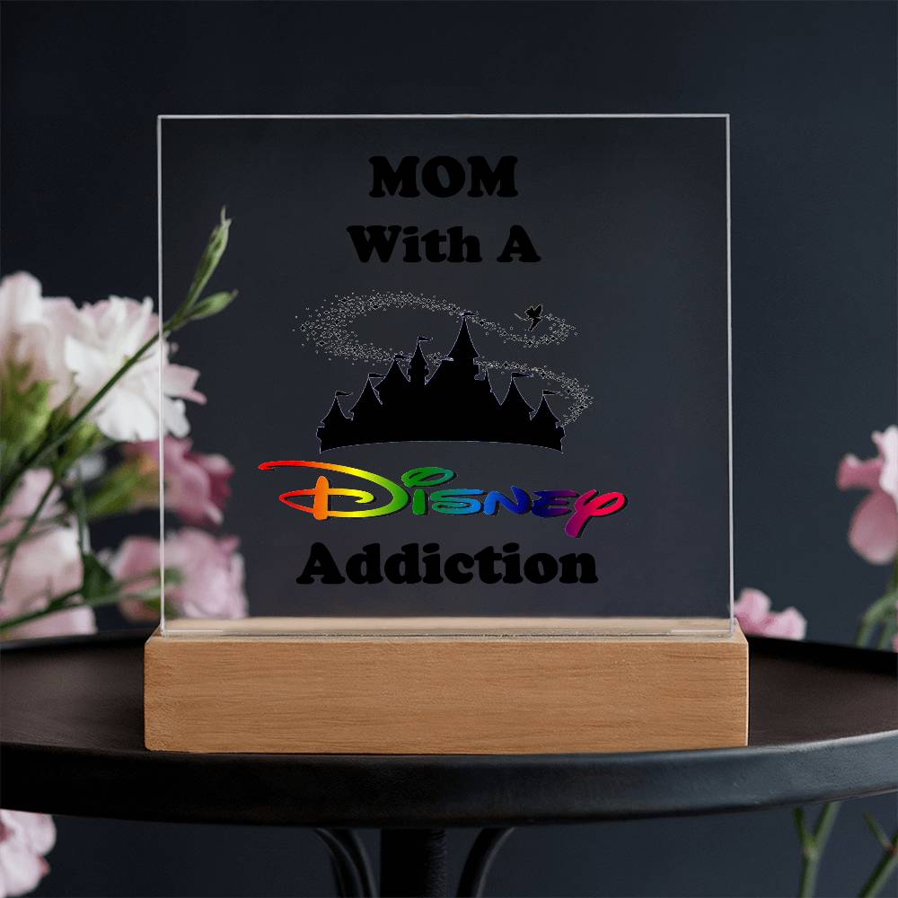 The "Disney-Obsessed Mom: Where Magic Meets Motherhood!" acrylic plaque by ShineOn Fulfillment features vibrant text that reads "MOM With A Disney Addiction," accompanied by a castle silhouette. This ideal gift for moms is elegantly displayed on a wooden base, resting atop a round black surface and surrounded by softly blurred flowers.