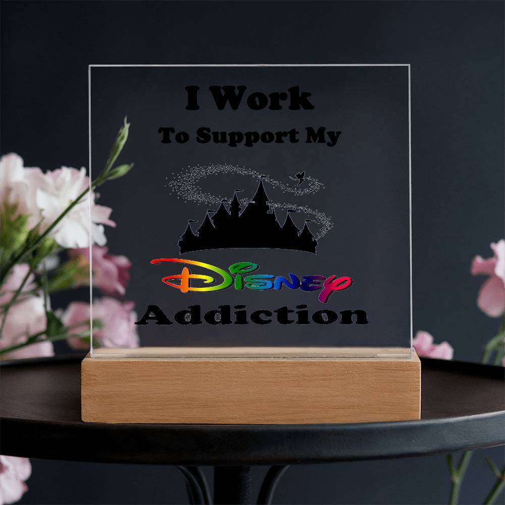 A Disney Obsessed: Where Magic Meets Everyday Life! acrylic plaque by ShineOn Fulfillment features a castle silhouette and vibrant "Disney" text, expressing the sentiment, "I Work To Support My Disney Addiction." This whimsical decor is beautifully enhanced by the soft blur of pink and white flowers in the background.