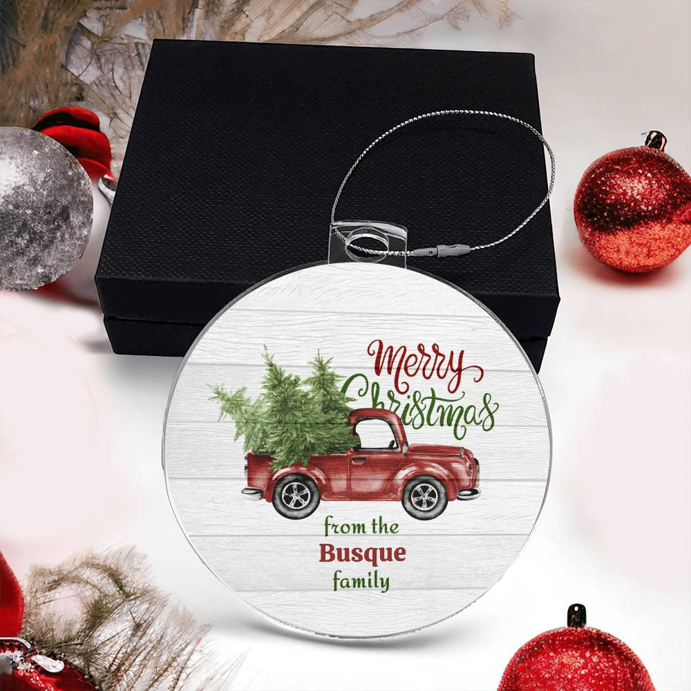 A festive Personalized Old Pickup Truck Ornament with Christmas Trees from AnywherePOD, showcasing a vintage pickup carrying a green Christmas tree. The phrase "Merry Christmas from the Busque family" is elegantly inscribed in red and green. It is displayed against a black gift box amidst holiday decorations.