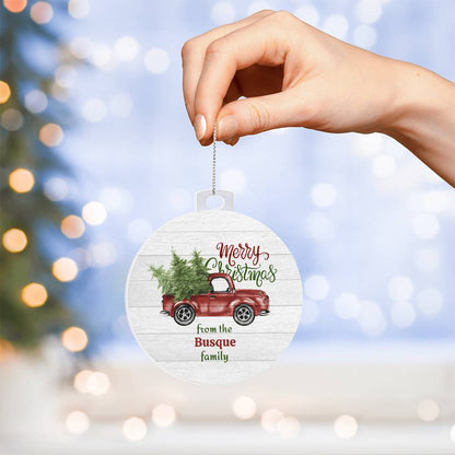 A hand holds the Personalized Old Pickup Truck Ornament with Christmas Trees from AnywherePOD, featuring a vintage pickup truck carrying two evergreen trees. The text reads "Merry Christmas from the Busque family," while a blurred Christmas tree with twinkling lights is in the background.