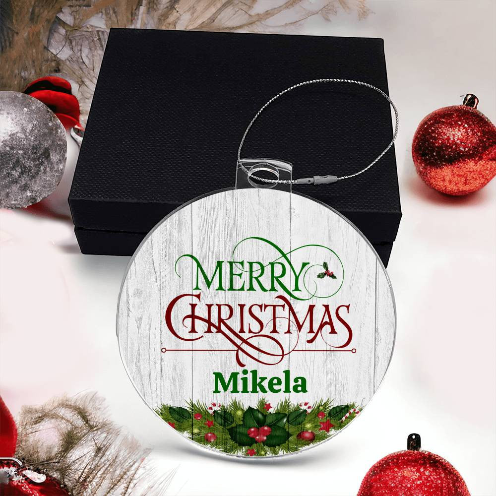 The Personalized Name Christmas Ornament by AnywherePOD, with the message "Merry Christmas Mikela" in festive red and green letters, elegantly adorns a black box and is surrounded by red and silver decorations. This acrylic ornament is enhanced with holly leaves and berries at the bottom, making it an ideal custom keepsake for your holiday tree.